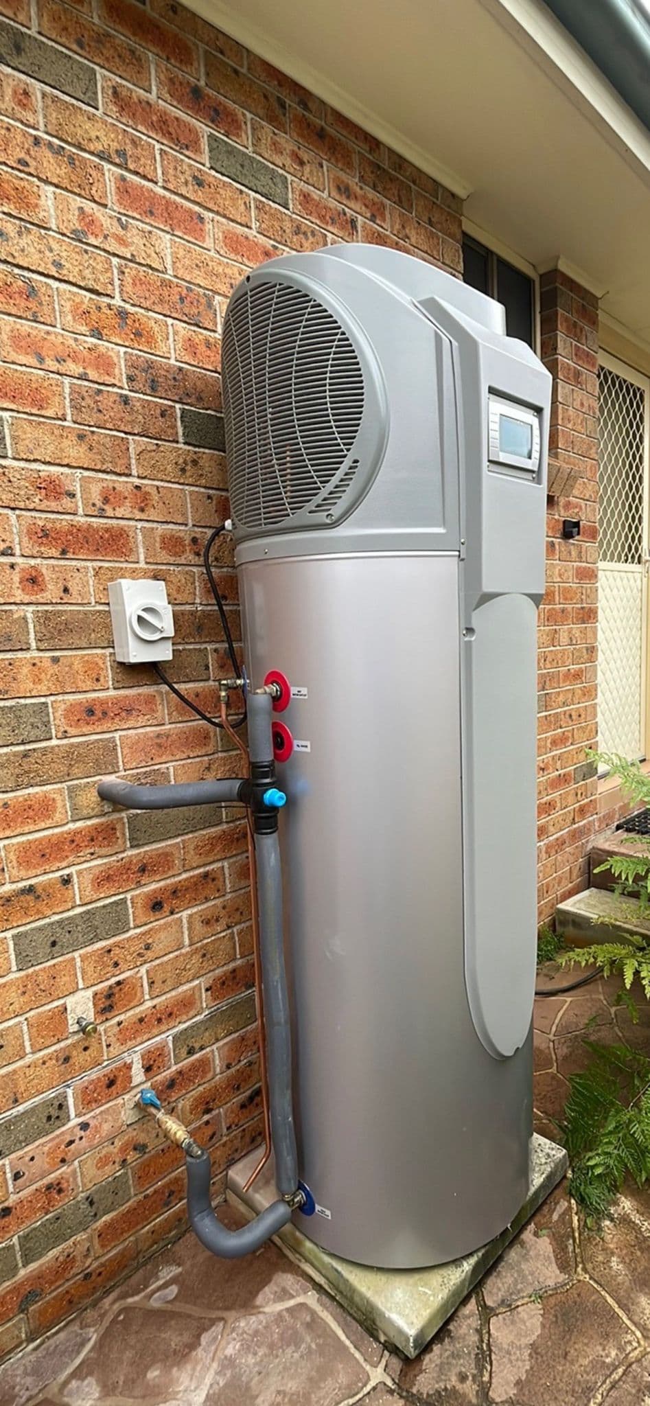 Heat-pump hot water unit and insulated connections beside a brick wall