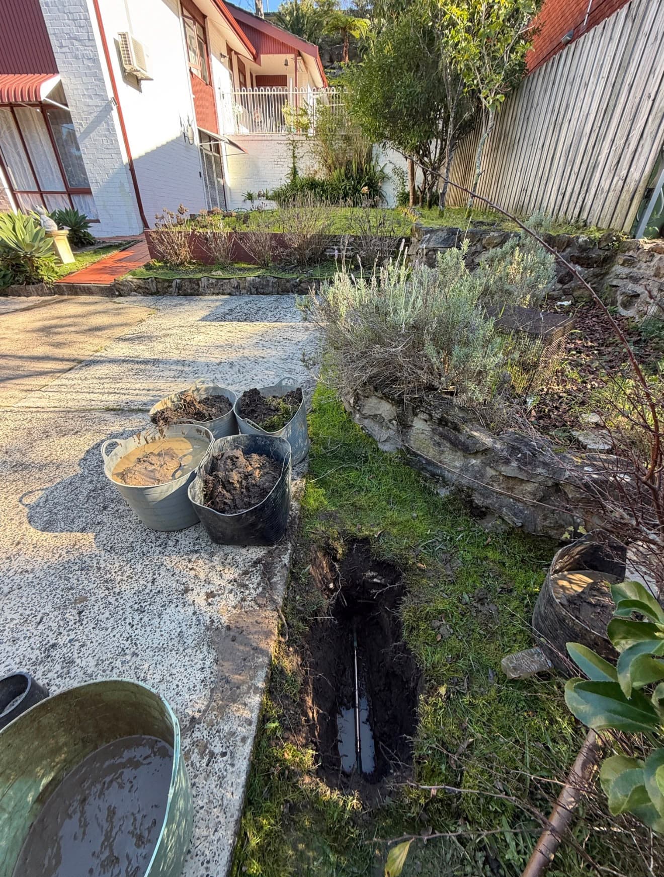 Narrow excavation beside a garden path with exposed pipework